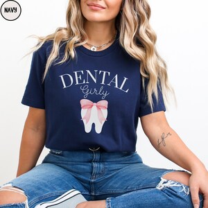 May include: A navy blue t-shirt with the text "Dental Girly" in white lettering. A white tooth with a pink bow is centered on the shirt.