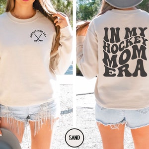May include: A beige sweatshirt with the text "In My Hockey Mom Era" in a retro font on the back. The front of the sweatshirt has a small graphic of hockey sticks with the text "Hockey Mom Era".