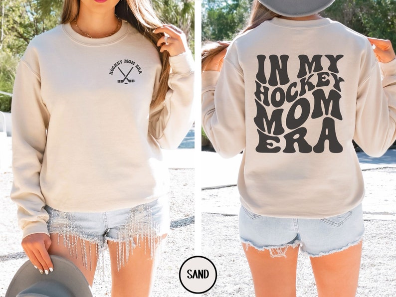 May include: A beige sweatshirt with the text "In My Hockey Mom Era" in a retro font on the back. The front of the sweatshirt has a small graphic of hockey sticks with the text "Hockey Mom Era".