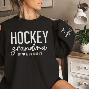 May include: Black sweatshirt with the text "HOCKEY grandma MY &hearts; IS ON THAT ICE" in white. The left sleeve has a white hockey stick and puck graphic with the text "Megan" in white.