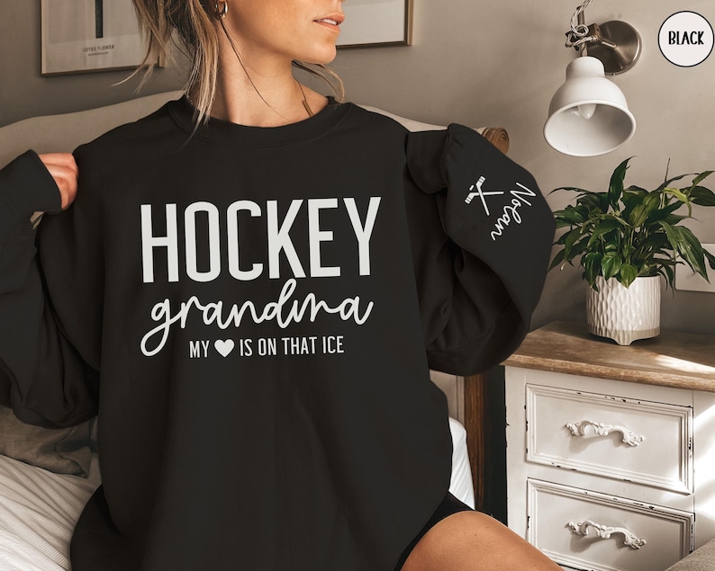 May include: Black sweatshirt with the text "HOCKEY grandma MY &hearts; IS ON THAT ICE" in white. The left sleeve has a white hockey stick and puck graphic with the text "Megan" in white.