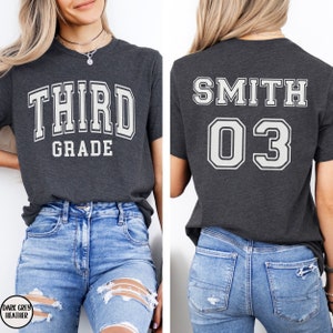 May include: Dark grey heather t-shirt with the words "THIRD GRADE" in white block letters on the front. The back of the shirt displays "SMITH" above the number "03" in white block letters. The shirt is paired with blue jeans.