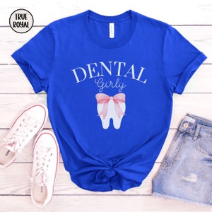 May include: A royal blue t-shirt with the text "Dental Girly" in white lettering. A white tooth with a pink bow is centered on the shirt.
