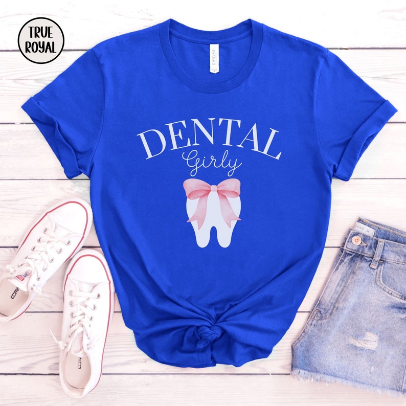 May include: A royal blue t-shirt with the text "Dental Girly" in white lettering. A white tooth with a pink bow is centered on the shirt.