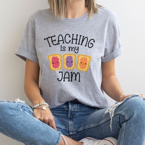 Peut inclure: Un t-shirt gris avec le texte "TEACHING is my JAM" et trois images de toasts de dessin anim&eacute; avec des confitures de diff&eacute;rentes couleurs.