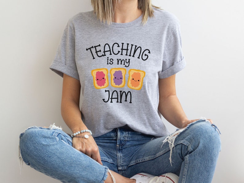 Peut inclure: Un t-shirt gris avec le texte "TEACHING is my JAM" et trois images de toasts de dessin anim&eacute; avec des confitures de diff&eacute;rentes couleurs.