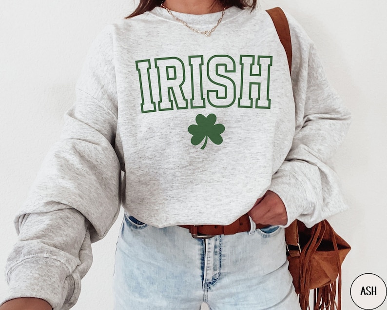 Op de afbeelding: Een grijs sweatshirt met het woord "IRISH" bedrukt in groene letters met een witte omtrek. Onder het woord staat een groene klaver.