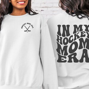 May include: White sweatshirt with a black graphic on the front that says "Hockey Mom Era" with crossed hockey sticks. The back of the sweatshirt has a black graphic that says "In My Hockey Mom Era" in a retro font.