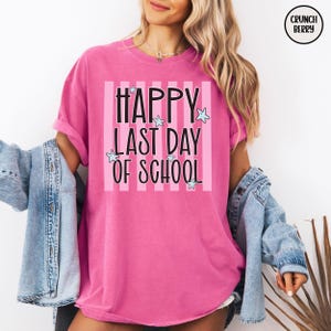 May include: A pink t-shirt with the text "HAPPY LAST DAY OF SCHOOL" in black, with light blue star accents. The text is centered on a pink and white striped background. The shirt has rolled-up sleeves.