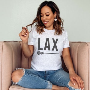 May include: A woman wearing a grey t-shirt with the text "LAX" and a lacrosse stick graphic. She is sitting on a pink couch.