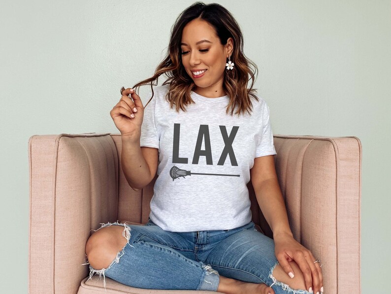 May include: A woman wearing a grey t-shirt with the text "LAX" and a lacrosse stick graphic. She is sitting on a pink couch.