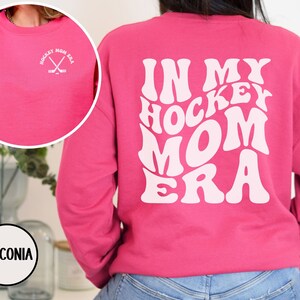 May include: Pink sweatshirt with the text "In My Hockey Mom Era" in white, retro-style lettering on the back. The front of the sweatshirt has a small white graphic of crossed hockey sticks with the text "Hockey Mom Era" in a circle.