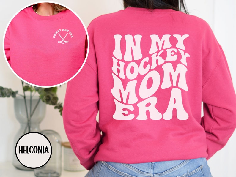 May include: Pink sweatshirt with the text "In My Hockey Mom Era" in white, retro-style lettering on the back. The front of the sweatshirt has a small white graphic of crossed hockey sticks with the text "Hockey Mom Era" in a circle.