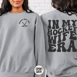Pu&ograve; includere: Felpa grigia con il testo "In My Hockey Wife Era" in un font retr&ograve; sul retro e il testo "Hockey Wife Era" con una grafica di bastone da hockey incrociato sul davanti.