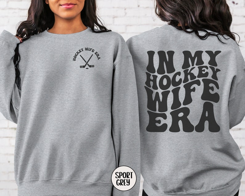 Pu&ograve; includere: Felpa grigia con il testo "In My Hockey Wife Era" in un font retr&ograve; sul retro e il testo "Hockey Wife Era" con una grafica di bastone da hockey incrociato sul davanti.