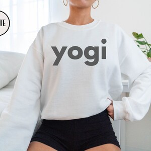 Może przedstawiać: Biała bluza z nadrukiem słowa "yogi" w kolorze czarnym z przodu.