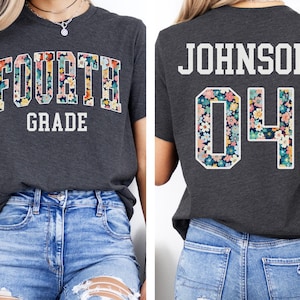 May include: Dark grey heather t-shirt with "FOURTH GRADE" in white text. The word "FOURTH" is filled with a floral pattern. The back of the shirt has the name "JOHNSON" and the number "04" in a floral pattern.