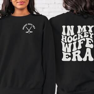 Pu&ograve; includere: Felpa nera con il testo "Hockey Wife Era" in un font retr&ograve; sul retro. La parte anteriore della felpa ha una piccola grafica di bastoni da hockey incrociati con il testo "Hockey Wife Era" in un font pi&ugrave; piccolo.