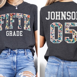 Op de afbeelding: Donkergrijze heather t-shirt met de witte tekst "FIFTH GRADE". De letters zijn gevuld met een bloemenpatroon in roze, geel en blauw. De achterkant van de shirt toont de naam "JOHNSON" en het nummer "05" met hetzelfde bloemendesign.