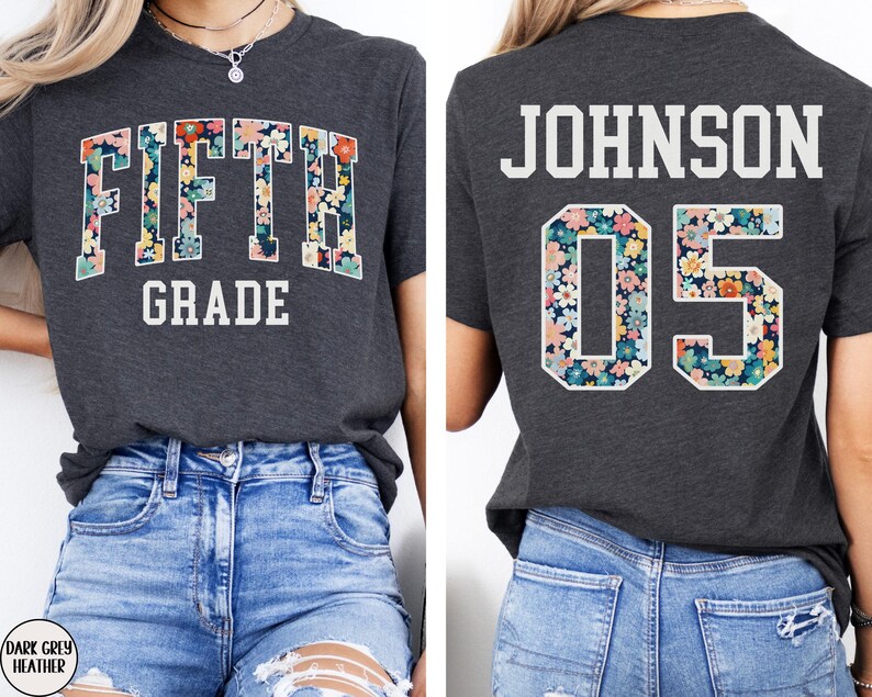Op de afbeelding: Donkergrijze heather t-shirt met de witte tekst "FIFTH GRADE". De letters zijn gevuld met een bloemenpatroon in roze, geel en blauw. De achterkant van de shirt toont de naam "JOHNSON" en het nummer "05" met hetzelfde bloemendesign.