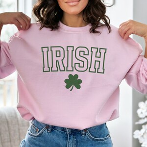 Op de afbeelding: Een lichtroze sweatshirt met het woord "IRISH" in groene blokletters en een groen klavertje vier in de onderkant.