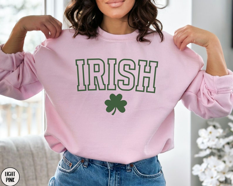 Op de afbeelding: Een lichtroze sweatshirt met het woord "IRISH" in groene blokletters en een groen klavertje vier in de onderkant.