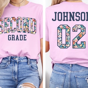 Puede incluir: Camiseta rosa claro con las palabras "SECOND GRADE" en un dise&ntilde;o floral en la parte delantera. La parte trasera de la camiseta tiene el nombre "JOHNSON" y el n&uacute;mero "02" en un dise&ntilde;o floral.