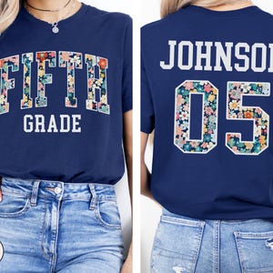 Op de afbeelding: Marineblauwe t-shirt met de tekst "FIFTH GRADE" in witte blokletters. Het woord "FIFTH" is gevuld met een bloemenpatroon. De achterkant toont de naam "JOHNSON" en het nummer "05" in een bloemenpatroon.