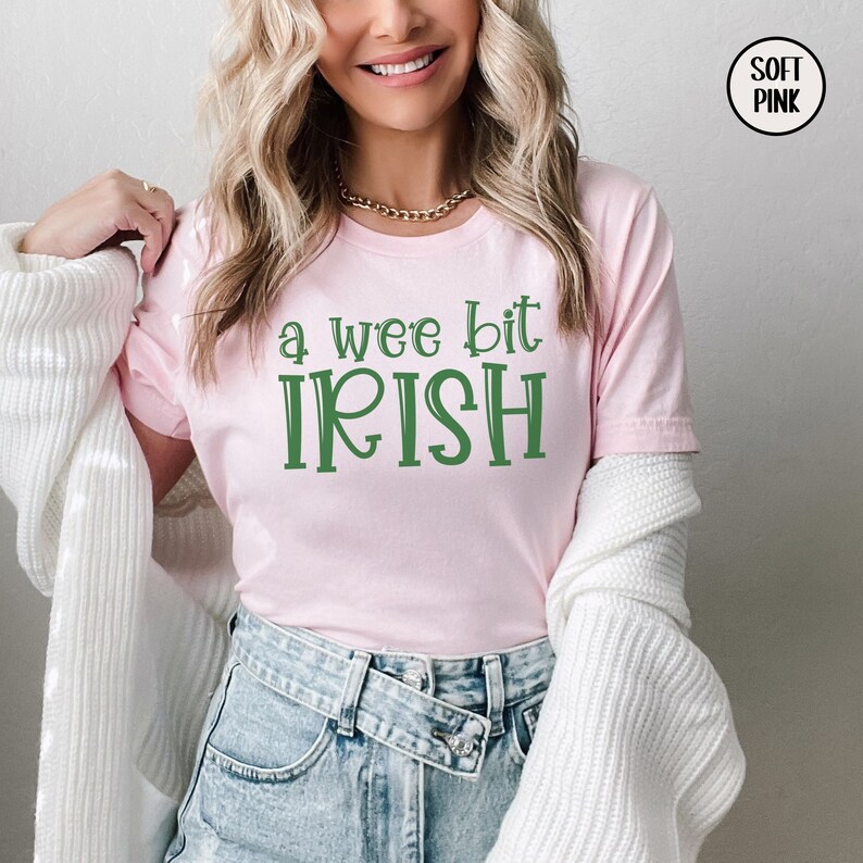 以下が含まれることがあります： 薄いピンク色のTシャツ。緑色の文字で「a wee bit IRISH」と書かれています。クルーネックの柔らかい素材です。聖パトリックの日のコレクションの一部です。