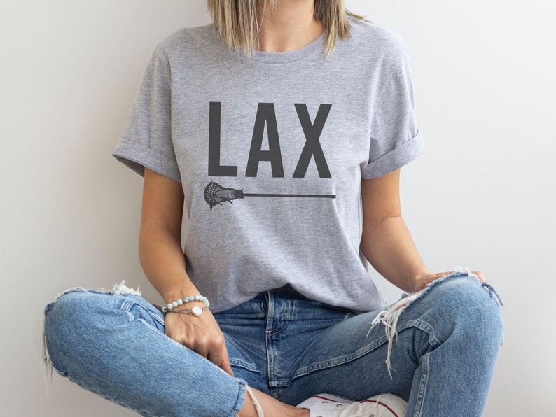 May include: A gray t-shirt with the word "LAX" printed in black on the front. A black lacrosse stick is printed below the word. The shirt is being worn by a person sitting down with their legs crossed.