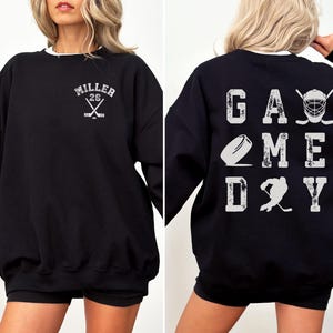 May include: Black sweatshirt with white text that says "Miller 26" and crossed hockey sticks on the front. The back of the sweatshirt says "Game Day" with a hockey puck, stick, and player graphic.