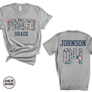 May include: A heather gray t-shirt with "FOURTH GRADE" in a floral design on the front. The back of the shirt displays "JOHNSON" and the number "04" in a floral pattern. The shirt is made of athletic heather material.