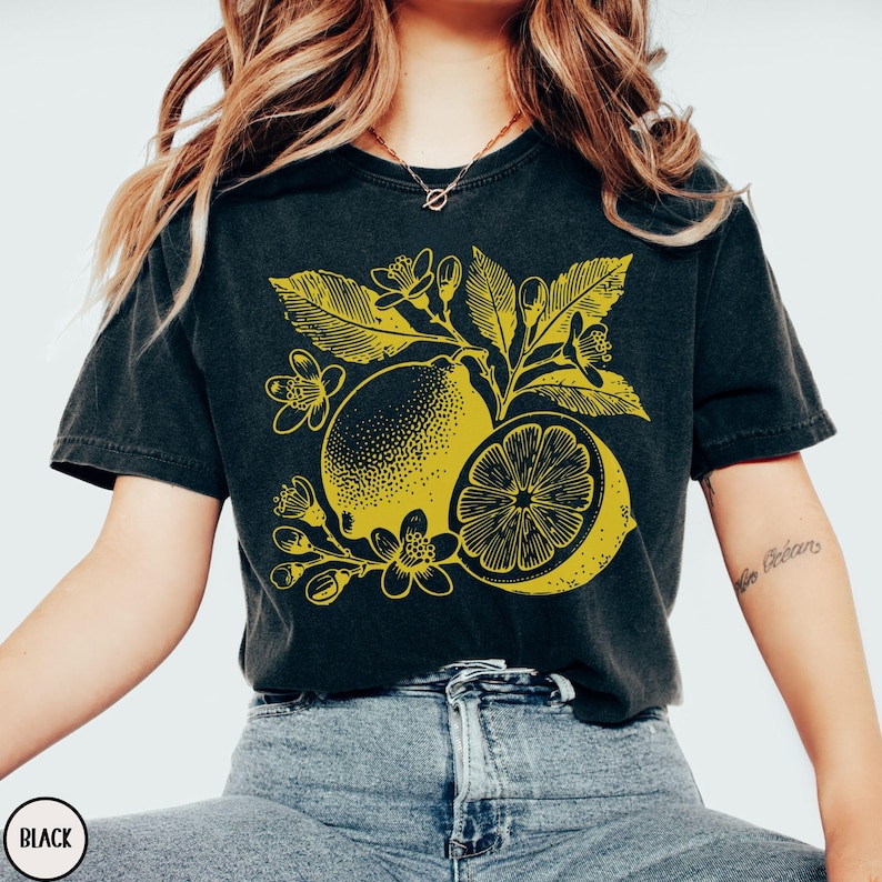 Op de afbeelding: Zwart T-shirt met een gele afbeelding van citroenen en bloemen. De afbeelding is gestileerd en heeft een vintage uitstraling.