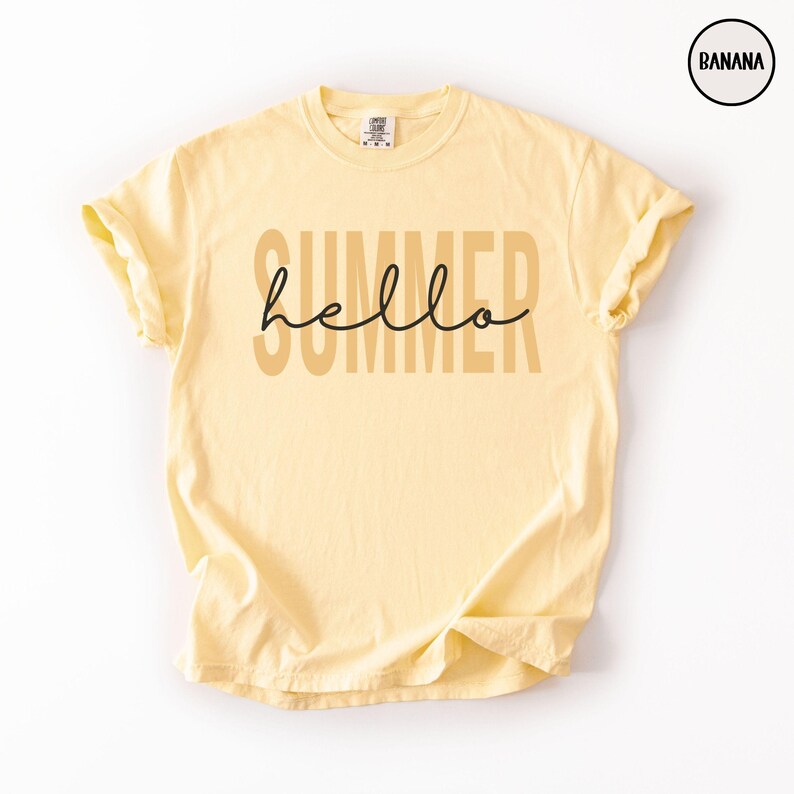 May include: A light yellow t-shirt with rolled-up sleeves. The word "SUMMER" is printed in a light brown color, with the word "hello" in a black cursive font overlaid on top of "SUMMER".
