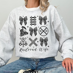 May include: Light gray sweatshirt with a railroad-themed design. The design includes black bows, a train, railroad crossing signs, and the text "Railroad Wife". The sweatshirt is made of a soft material.