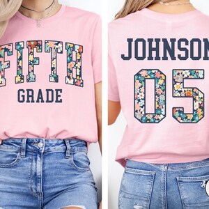 Op de afbeelding: Een lichtroze t-shirt met de woorden "FIFTH GRADE" in een bloemenpatroon. De achterkant van de shirt heeft de naam "JOHNSON" en het nummer "05" in een bloemenpatroon. Het shirt is gemaakt van een zacht materiaal.