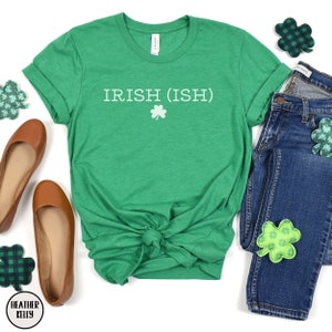 May include: Green t-shirt with the text "IRISH (ISH)" in white with a white shamrock underneath. The shirt is tied in a knot in the front and is laid out on a white background with a pair of brown flats, a pair of blue jeans, and green shamrock patches.