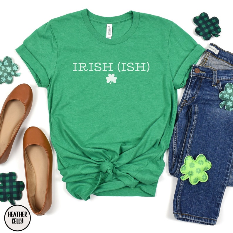 May include: Green t-shirt with the text "IRISH (ISH)" in white with a white shamrock underneath. The shirt is tied in a knot in the front and is laid out on a white background with a pair of brown flats, a pair of blue jeans, and green shamrock patches.
