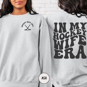 Pu&ograve; includere: Una felpa grigia con il testo "In My Hockey Wife Era" in un font retr&ograve; sul retro e una piccola grafica di bastoni da hockey incrociati con il testo "Hockey Wife Era" sul davanti.