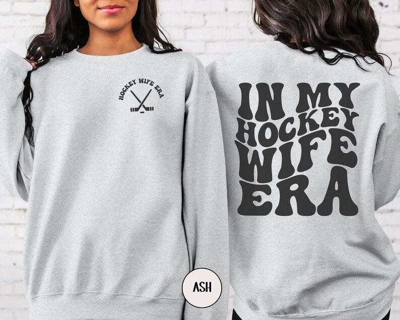 Pu&ograve; includere: Una felpa grigia con il testo "In My Hockey Wife Era" in un font retr&ograve; sul retro e una piccola grafica di bastoni da hockey incrociati con il testo "Hockey Wife Era" sul davanti.