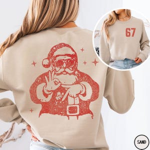May include: Sand-colored sweatshirt featuring a distressed red graphic of Santa Claus making a hand gesture. The graphic includes Santa's hat, sunglasses, and beard. A smaller image shows the sweatshirt with the number "67" in red. The sweatshirt is a casual, festive garment.