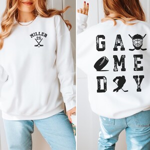 May include: White sweatshirt with a black hockey-themed design on the front and back. The front design says "Miller 26" with crossed hockey sticks. The back design says "GAME DAY" with a hockey puck, a hockey player, and a hockey mask.