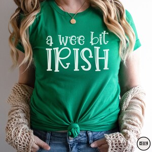 以下が含まれることがあります： 白い文字で「a wee bit IRISH」と書かれたエメラルドグリーンのTシャツ。 シャツはウエストで結ばれています。 ベージュのカーディガンが肩にかけられています。 ゴールドのネックレスが見えます。