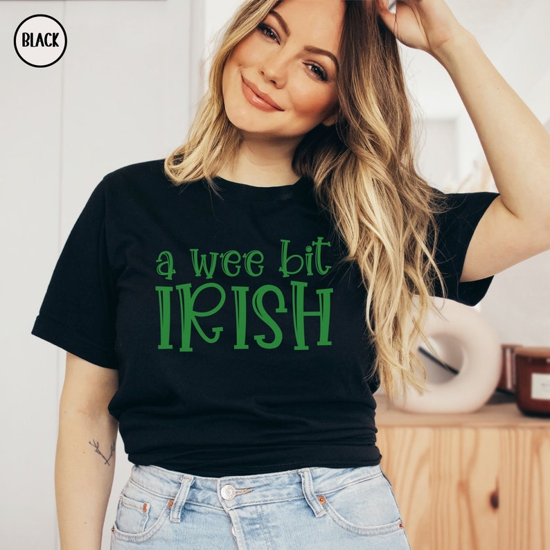 以下が含まれることがあります： 緑色の「a wee bit IRISH」の文字が入った黒いTシャツ。Tシャツはクルーネックで半袖です。このフレーズは、アイルランド文化や聖パトリックの日の祝賀にふさわしい装飾的なフォントで書かれています。