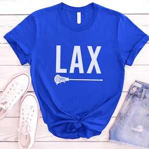 May include: A royal blue t-shirt with the word "LAX" printed in white on the front. A white lacrosse stick is printed below the word. The t-shirt is laid flat on a white wooden surface.