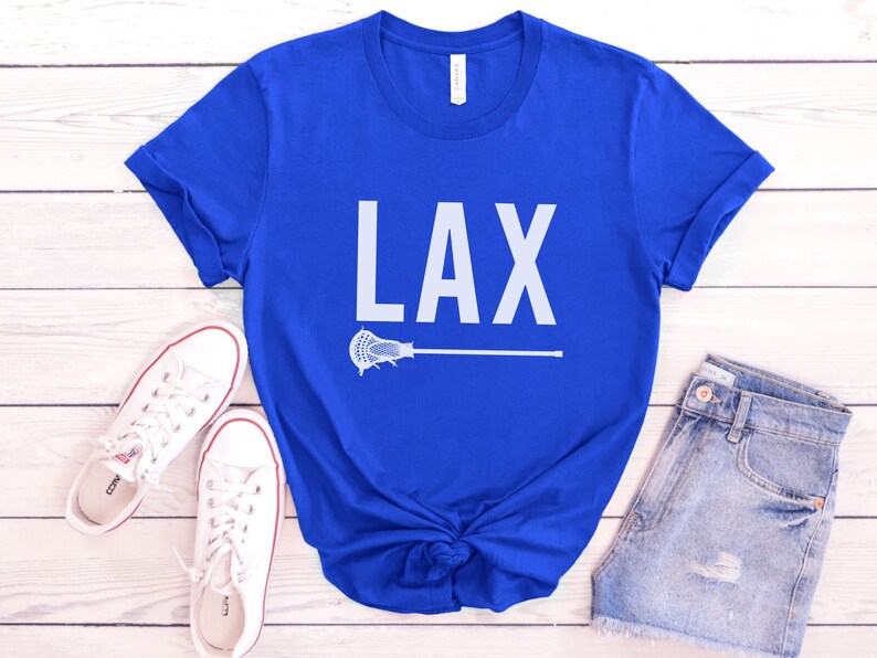 May include: A royal blue t-shirt with the word "LAX" printed in white on the front. A white lacrosse stick is printed below the word. The t-shirt is laid flat on a white wooden surface.
