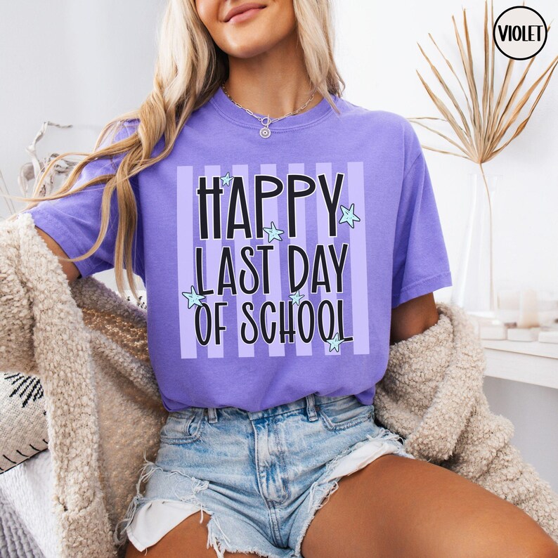 May include: A lilac-colored t-shirt with the text "HAPPY LAST DAY OF SCHOOL" in black, accented with light blue stars. The text is set against a backdrop of vertical stripes. The shirt is part of a clothing collection.
