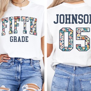 Op de afbeelding: Wit T-shirt met "FIFTH GRADE" in een bloemenpatroon op de voorkant. De achterkant van het shirt toont "JOHNSON 05" met een bloemenpatroon. Een casual T-shirt, geschikt voor school of dagelijks gebruik.