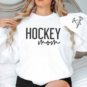May include: White sweatshirt with the text "HOCKEY mom" and a hockey stick graphic on the sleeve. The text "HOCKEY mom" is in black and the graphic is in black and white.