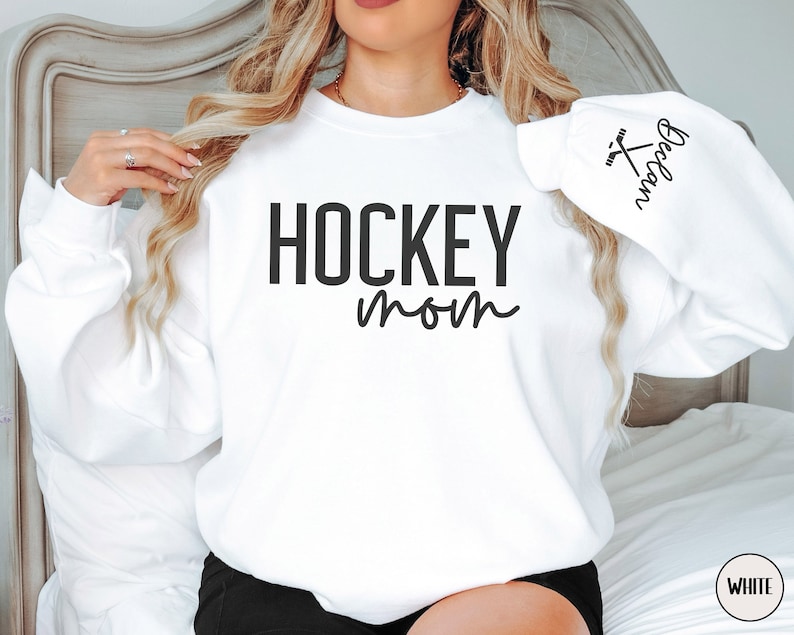May include: White sweatshirt with the text "HOCKEY mom" and a hockey stick graphic on the sleeve. The text "HOCKEY mom" is in black and the graphic is in black and white.